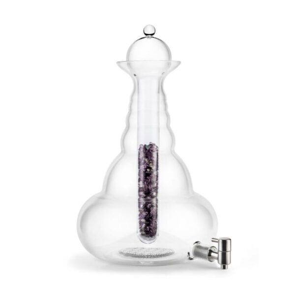 Eine große, bauchige Glaskaraffe mit Deckel und Zapfhahn; in der Mitte befindet sich ein Glaszylinder mit violetten Steinen.