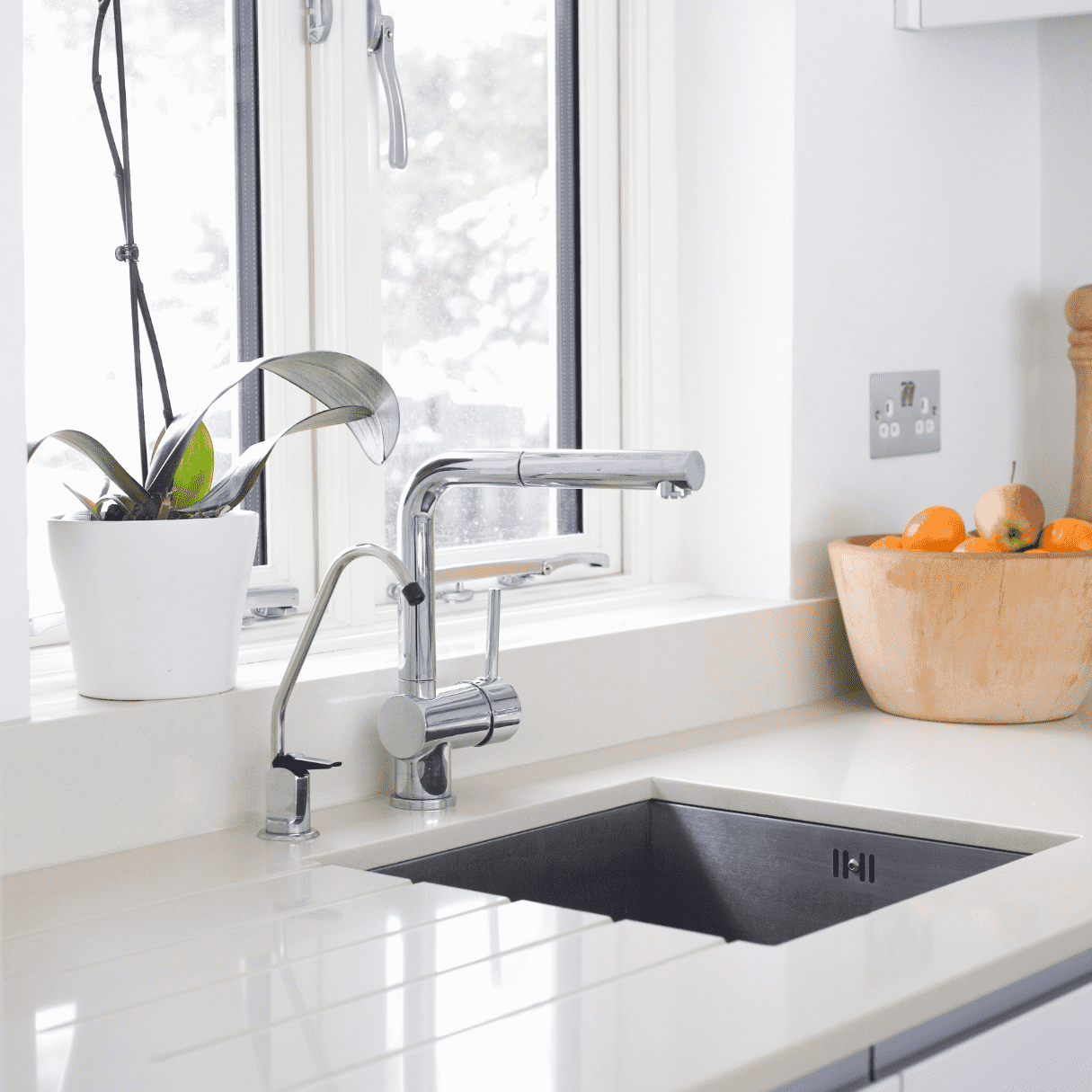 Eine moderne Küchenspüle mit glänzendem Wasserhahn, daneben ein Seifenspender, ein Blumentopf mit grüner Pflanze und eine Holzschale mit Orangen und Äpfeln auf heller Arbeitsplatte vor einem Fenster.
