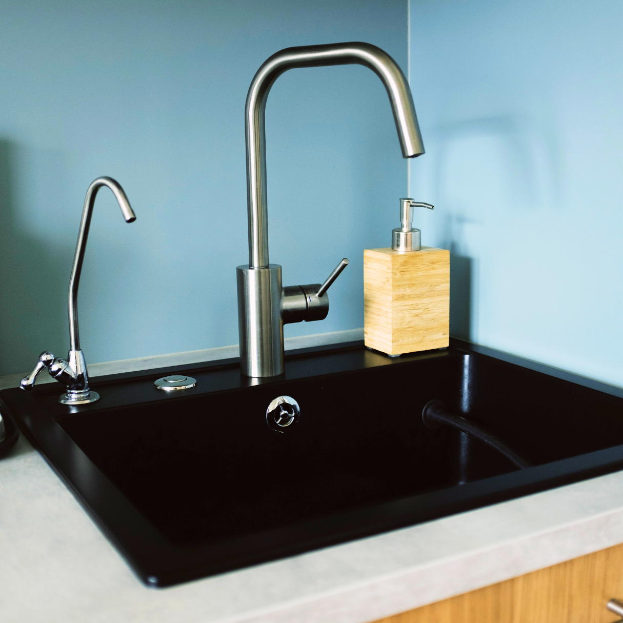 Ein modernes Spülbecken mit schwarzer Oberfläche, Edelstahlarmaturen, Seifenspender aus Holz und hellem Arbeitsplattenrand.