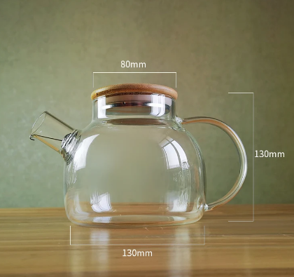 Eine transparente Teekanne aus Borosilikatglas mit Holzdeckel, seitlichem Glashenkel und Ausguss steht auf einer Holzoberfläche. Maße sind 130 mm breit, 130 mm hoch, Deckel 80 mm.