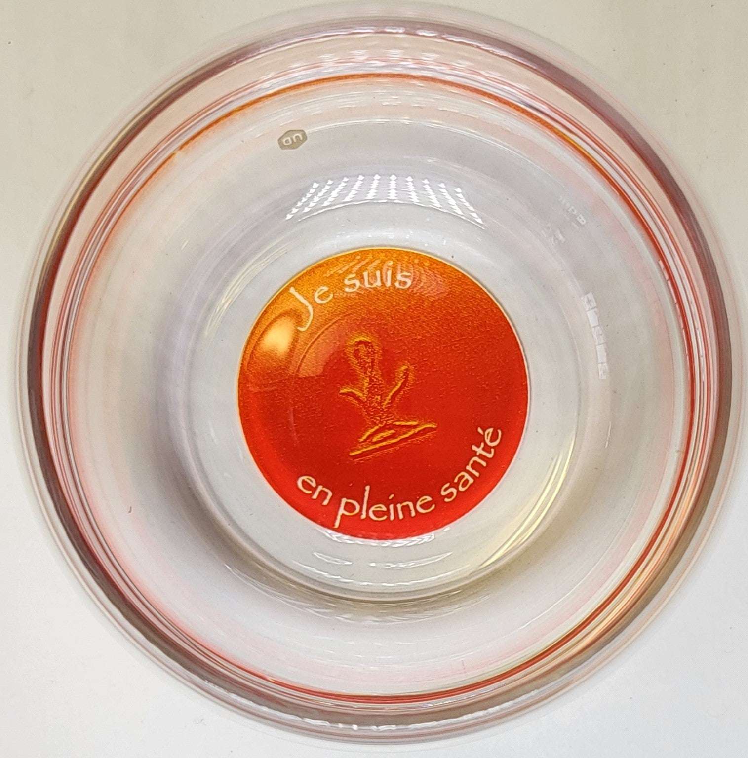 Ein rundes, durchsichtiges Glas mit orangerotem Boden, auf dem „Je suis en pleine santé“ und ein einfaches Pflanzensymbol stehen.