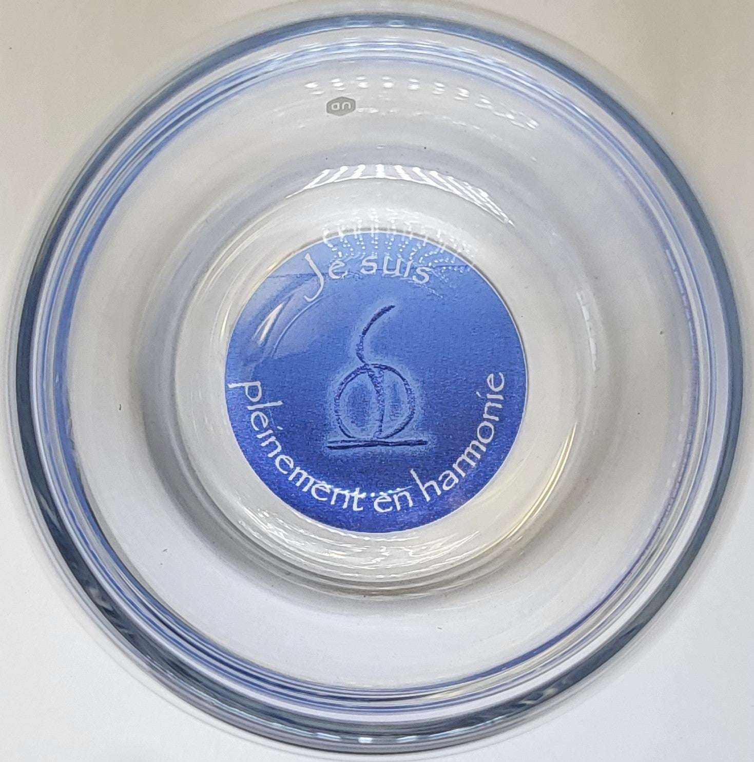 Ein runder, transparenter Glasbecher mit einem blauen Kreis am Boden. Auf dem Kreis steht in Weiß "Je suis pleinement en harmonie" und zeigt ein abstraktes Symbol in der Mitte. Das Glas hat eine dezente, leicht blaue Schattierung und liegt auf einer hellen Oberfläche. Die Aufschrift und das Symbol sind zentral positioniert.