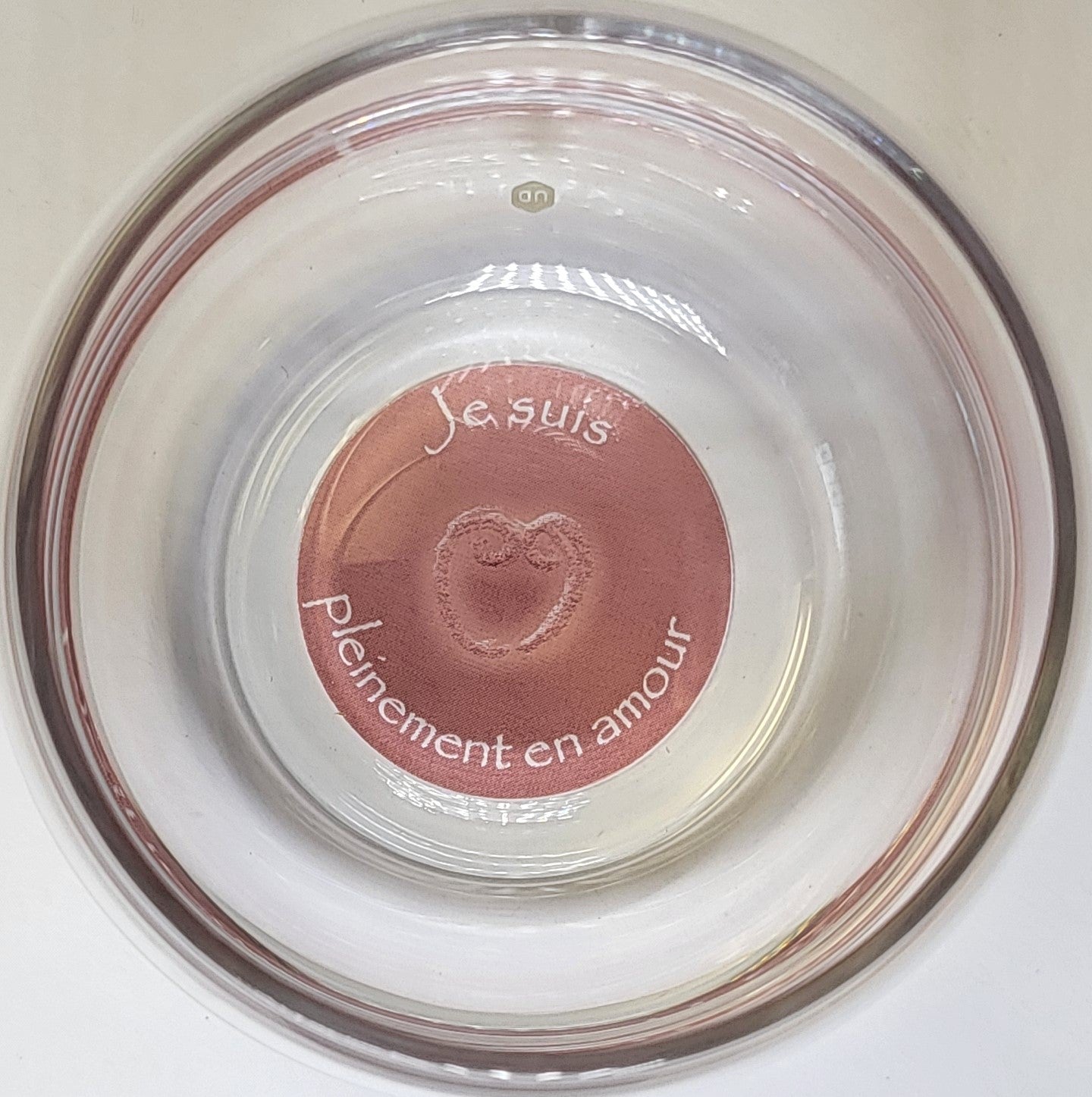 Ein transparenter Glasbecher von oben fotografiert, Boden mit rosa Kreis, Herzsymbol und französischer Aufschrift “Je suis pleinement en amour”.