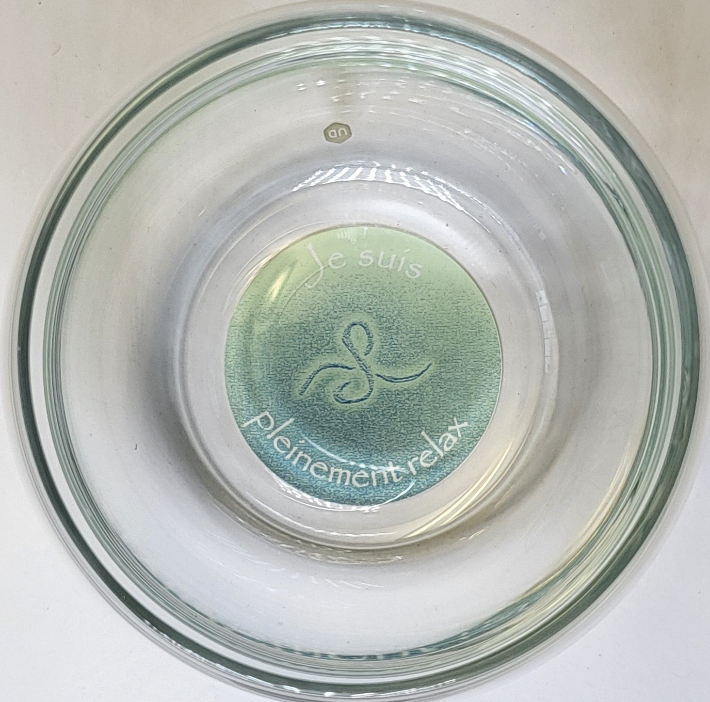 Ein durchsichtiges Glas mit rundem Boden, innen graviertem Schriftzug „Je suis Pleinement relax“ und stilisiertem Symbol.