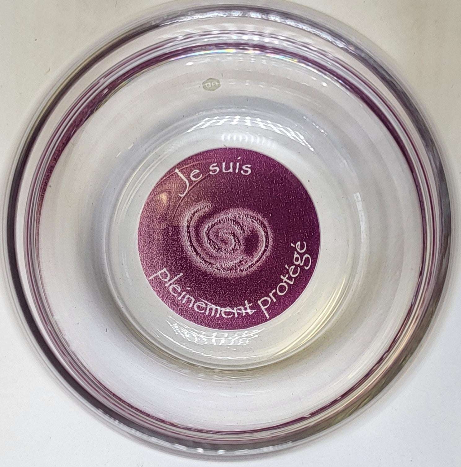 Ein klarer Mythos Becher mit violettem, rundem Boden, auf dem in Weiß „Je suis pleinement protégé“ und ein Spiralmotiv stehen.