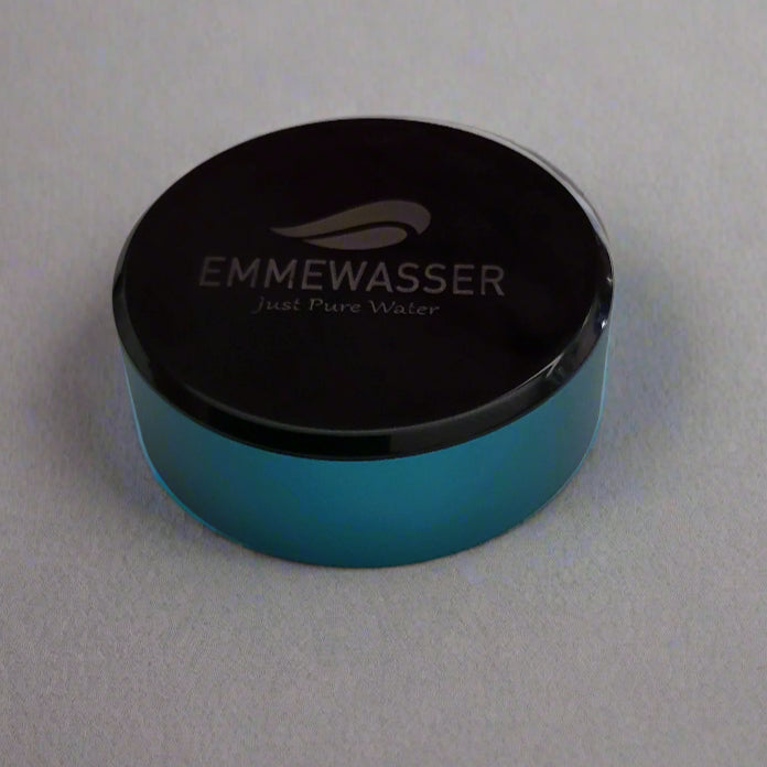 Eine runde, flache Komponente mit transparent-türkisem Rand und schwarzer Oberseite, Aufdruck „EMMEWASSER Just Pure Water“.