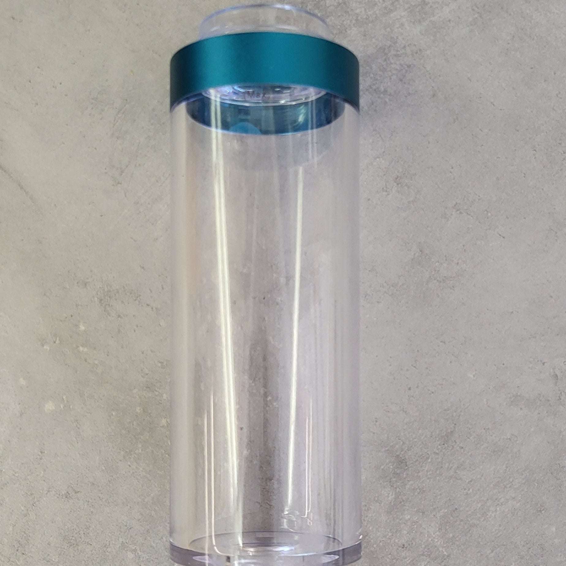 Ein zylindrischer transparenter Kunststoffbehälter mit einem türkisfarbenen Deckel, stehend auf einer grauen Oberfläche.