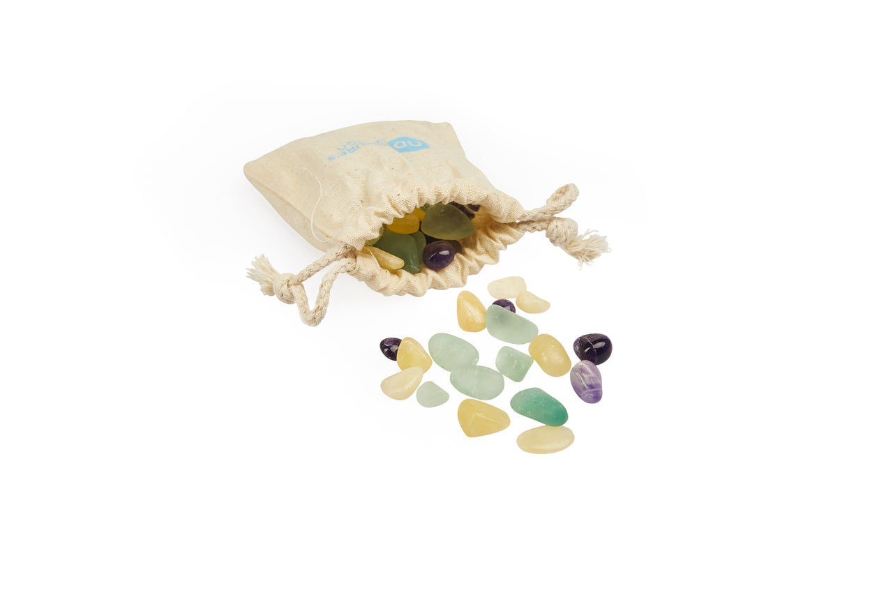 Ein cremefarbener Stoffbeutel mit Zugband, gefüllt mit bunten Edelsteinen in Gelb, Grün und Violett, einige herausgefallen.