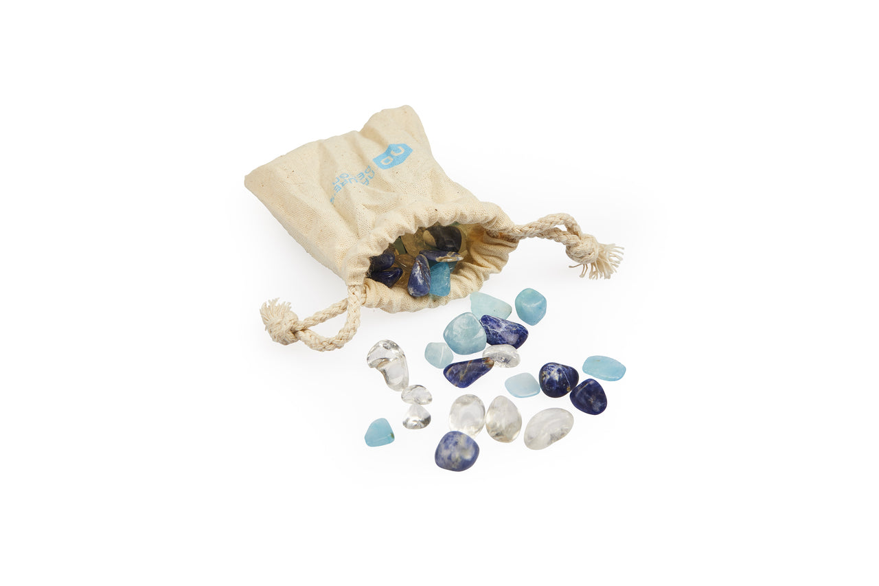 Ein beiger Stoffbeutel mit Kordel liegt auf weißem Hintergrund, teilweise geöffnet, und enthält verschiedene kleine, glatte Edelsteine. Die Edelsteine sind teils aus dem Beutel herausgefallen und breiten sich aus. Die Steine sind unterschiedlich groß und haben verschiedene Farben: Es gibt durchsichtige, hellblaue und dunkelblaue Steine. Der Beutel wirkt aus Leinstoff und hat einen dezenten blauen Aufdruck. Die Edelsteine sehen poliert und glänzend aus, teilweise mit weißen und blauen Maserungen.