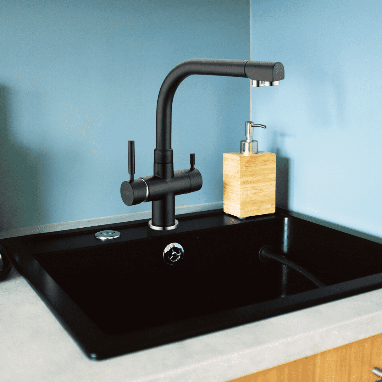Eine moderne schwarze Küchenspüle mit passender Armatur vor hellblauer Wand, daneben ein Seifenspender aus Holzoptik.