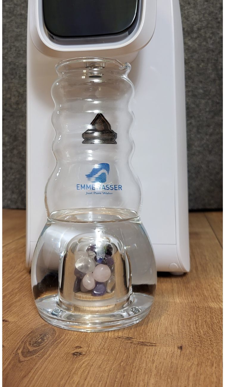 Eine Cadus Karaffe 1 Liter aus Glas mit gewellter Form, gefüllt mit Wasser und Edelsteinen im Bodengefäß, steht auf Holz.