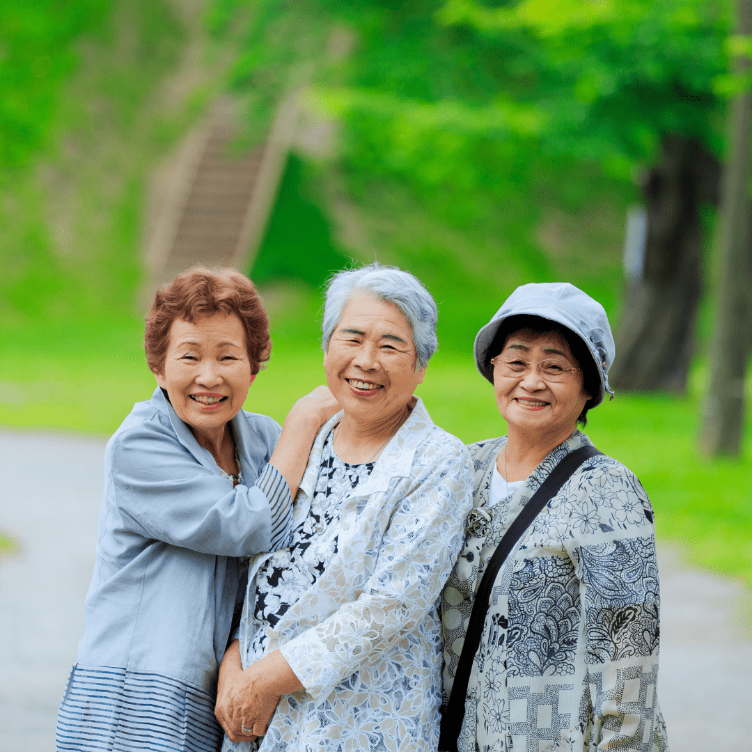 Drei ältere Frauen stehen dicht beieinander draußen, lächeln freundlich, tragen helle Jacken und eine Sonnenhut, im Park.