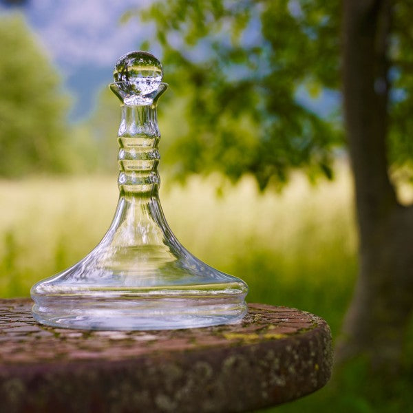 Eine elegante Rubellum Weinkaraffe aus klarem Glas, 1 Liter, mit rundem Stöpsel, steht auf einem rustikalen Holztisch im Freien.