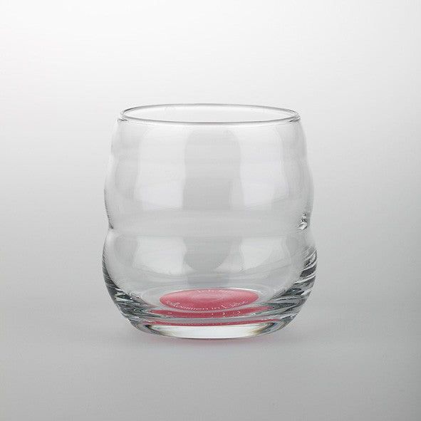 Ein klares, rundes Mythos Becher Glas mit welliger Außenform, rosa Akzent und Affirmation am Boden, auf weißem Hintergrund.