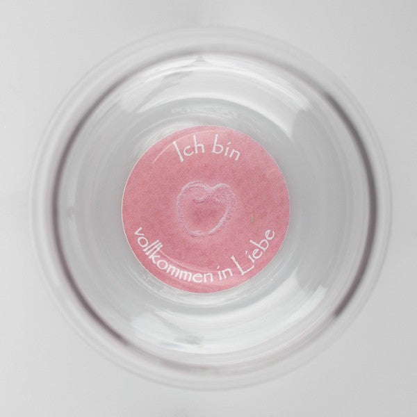 Ein transparenter Mythos Becher mit einem rosafarbenen Boden, darauf ein Herzsymbol und der Schriftzug „Ich bin vollkommen in Liebe“.