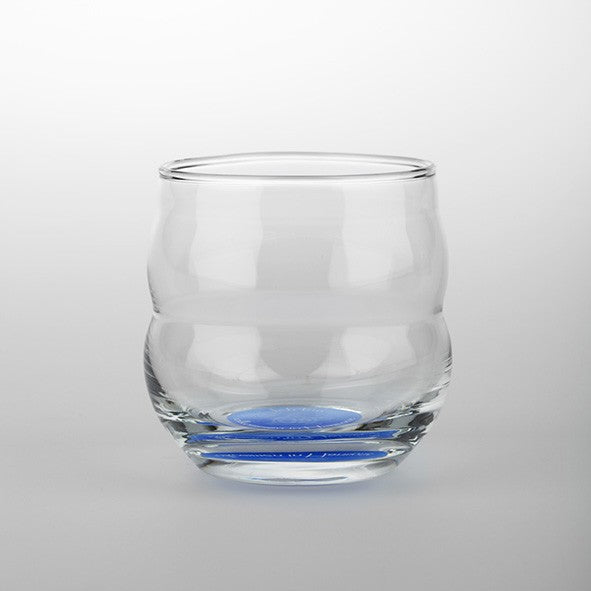 Ein transparentes Glas mit wellenförmiger Struktur und runden Formen, dicker Boden mit blauem Akzent, minimalistisches Design.