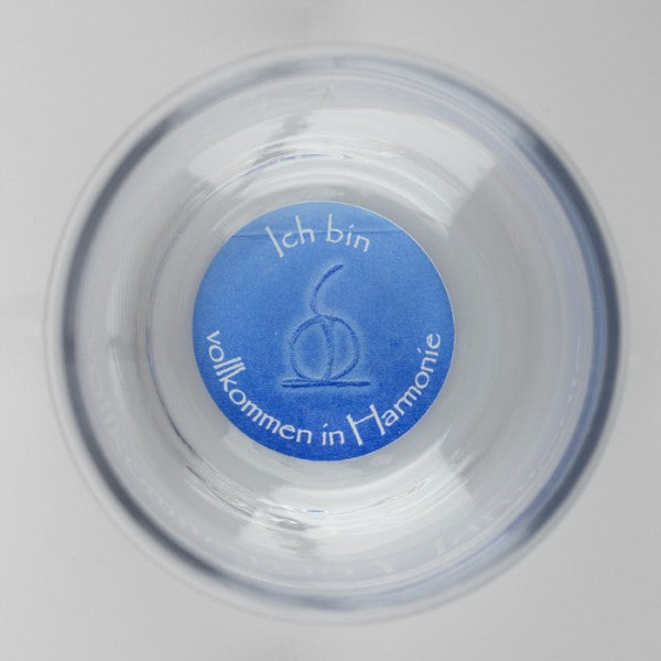 Ein transparenter Glasbecher, von oben fotografiert, mit einem blauen Kreis am Boden, der die weiße Aufschrift „Ich bin vollkommen in Harmonie“ und ein stilisiertes, eingraviertes Symbol zeigt.