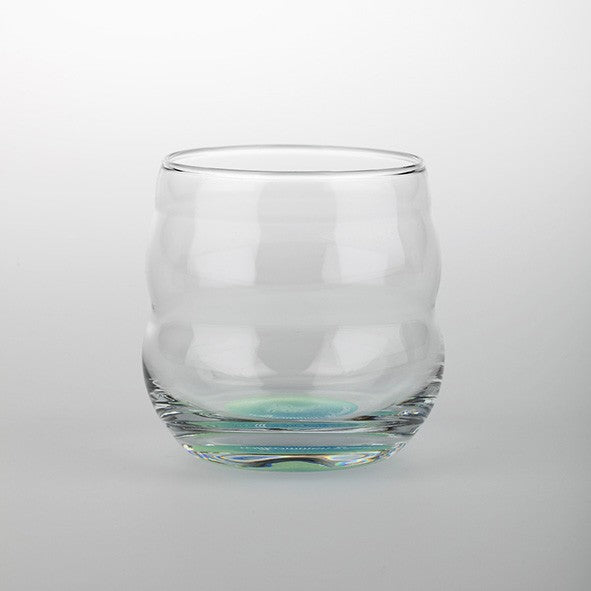 Ein rundes, klares Mythos Becher Glas mit welligen Konturen und dickem Boden, unten schimmernde grüne Akzente.