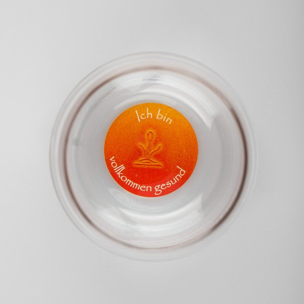 Ein durchsichtiger Mythos Becher mit orangefarbenem Boden, Aufschrift „Ich bin vollkommen gesund“ und stilisiertem Meditationssymbol.