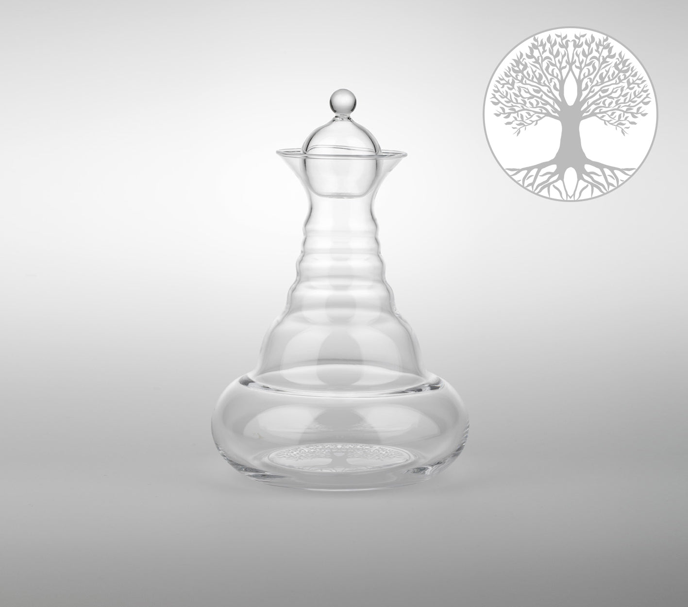 Eine transparente, bauchige Glaskaraffe mit gewelltem Hals und rundem Glasdeckel; daneben ein stilisierter Lebensbaum.