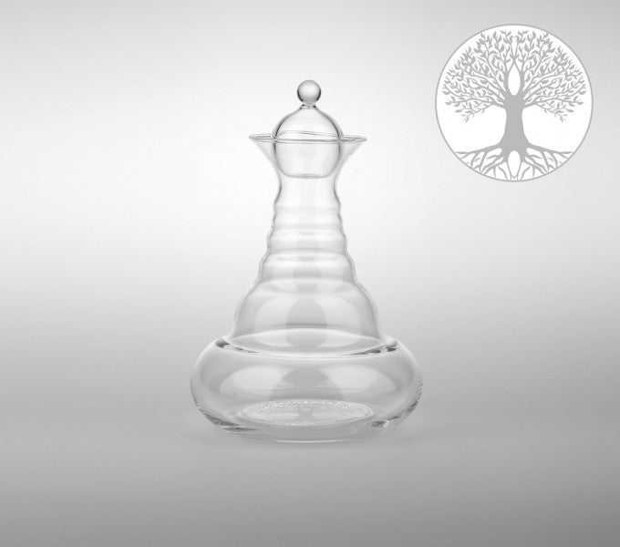 Das Bild zeigt eine klare, bauchige Glaskaraffe mit rundem Stopfen, daneben ein stilisierter Baum des Lebens als Symbol.