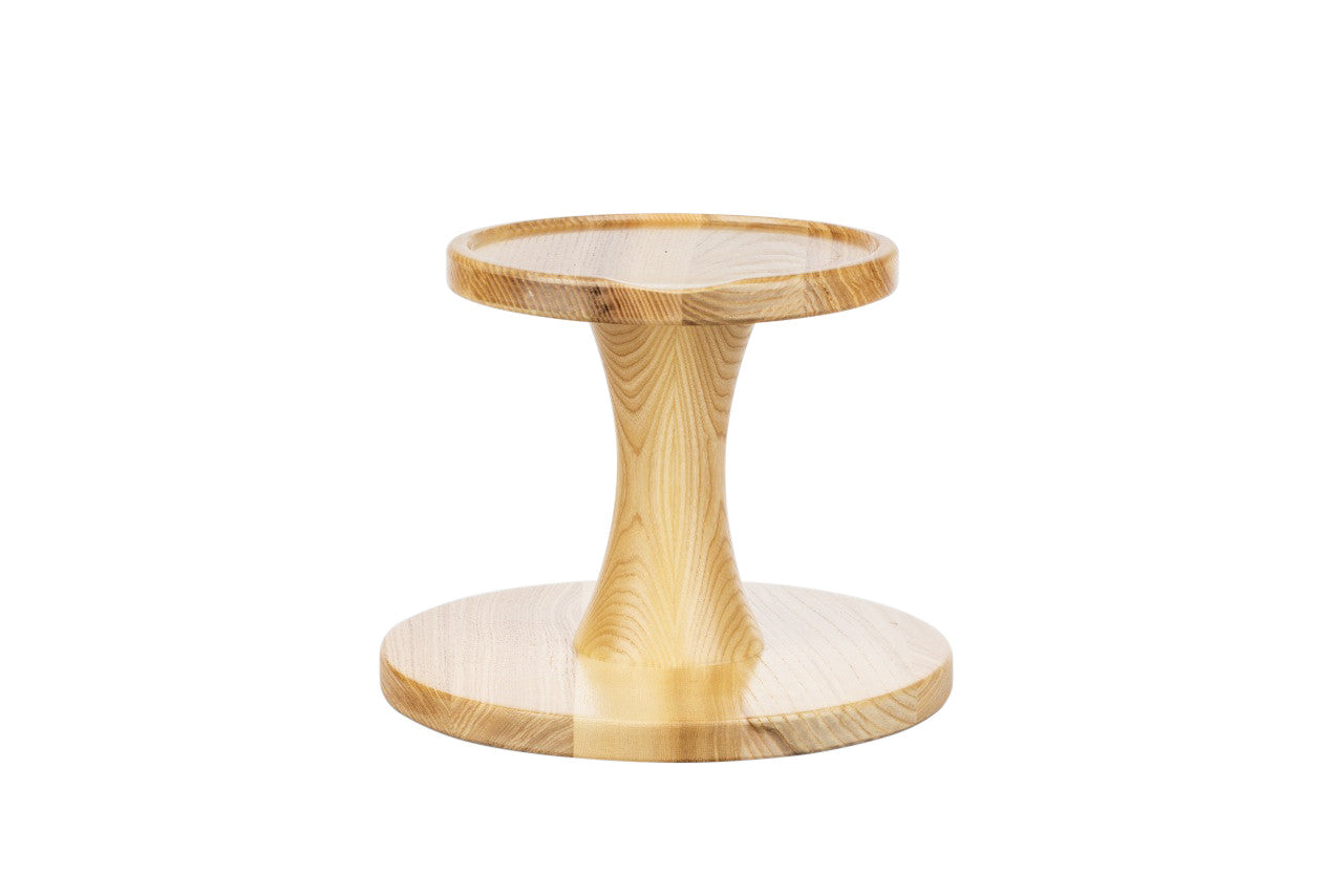 Zu sehen ist ein Unterbau aus hellem Eschenholz mit rundem Fuß und runder Ablage, dazwischen ein eleganter, konisch geformter Mittelsteg.