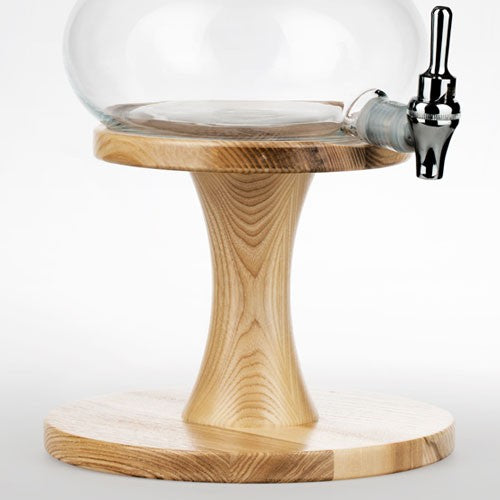 Ein heller, elegant geschwungener Unterbau aus Eschenholz hält eine große Glas-Karaffe mit silbernem Auslaufhahn.