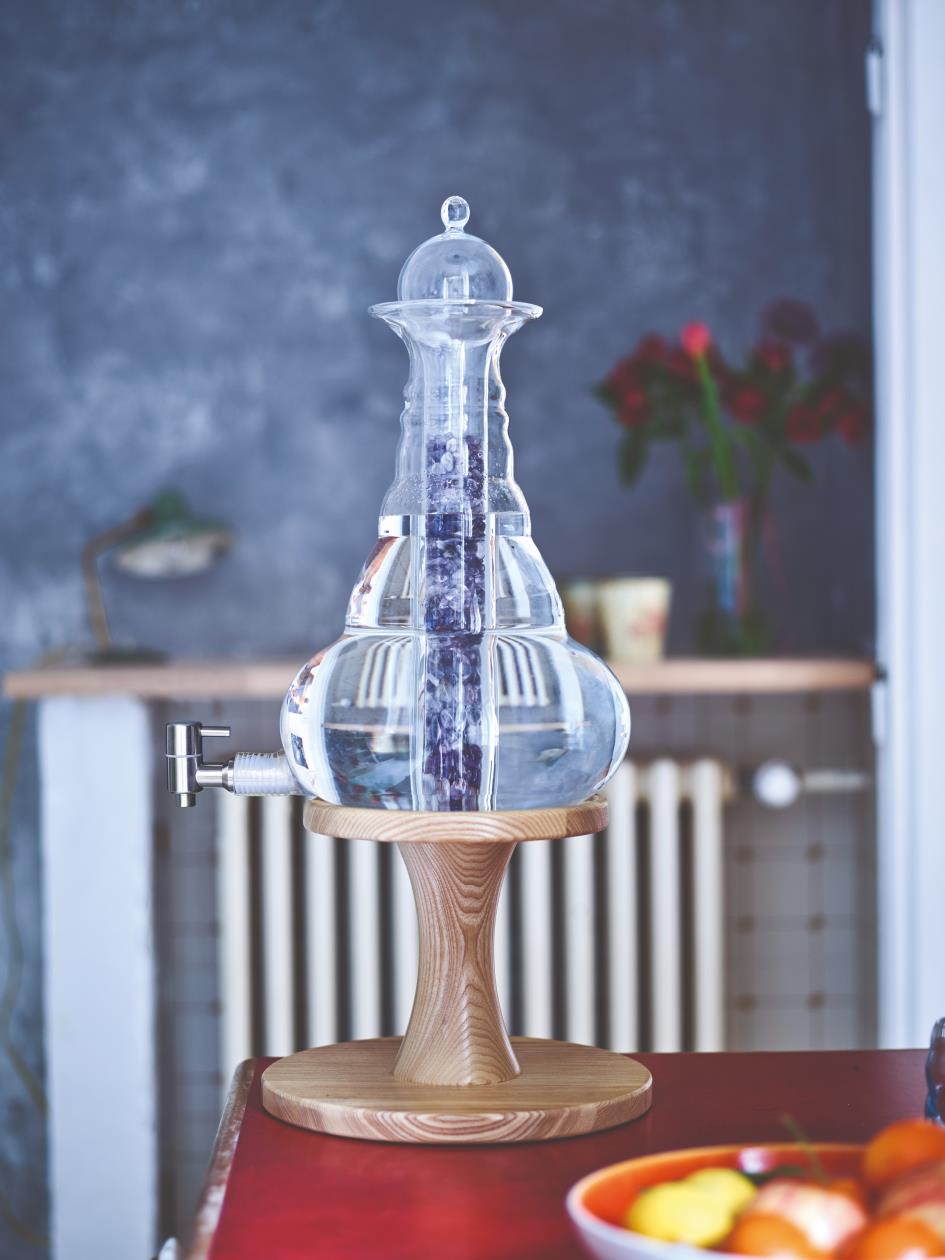 Auf einem hölzernen Unterbau aus Esche steht eine große, bauchige Glaskaraffe mit Deckel und Zapfhahn. Im Inneren befindet sich ein zylindrischer Einsatz mit hellvioletten Kristallen. Im Vordergrund ist eine rote Tischplatte mit einer orangefarbenen Schale, die Zitronen und Tomaten enthält. Im Hintergrund sind eine Heizung, eine Arbeitsplatte und eine Vase mit roten Blumen zu sehen. Die Umgebung wirkt wie eine gemütliche, modern eingerichtete Küche oder Esszimmer.