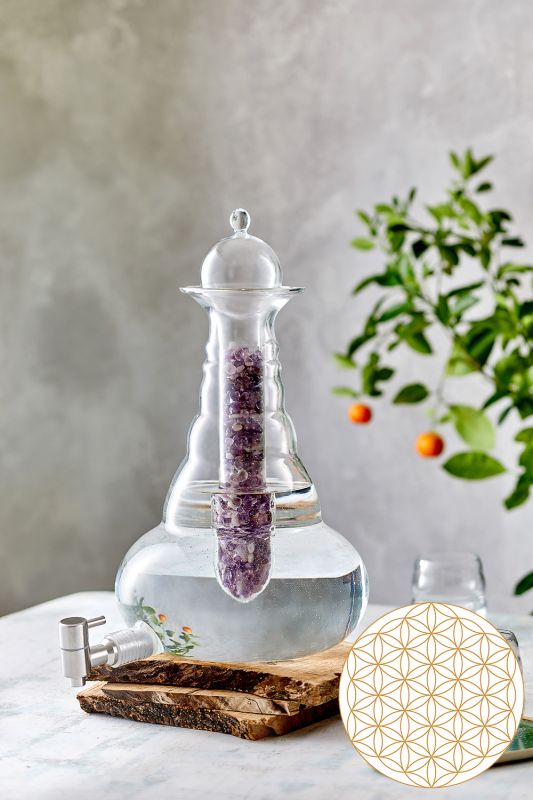 Eine große, bauchige Alladin Karaffe aus Glas mit 5 Litern Fassungsvermögen, gefüllt mit Wasser, integriertem Edelsteinmodul mit lilafarbenen Steinen, Holzuntersetzer, silberner Auslaufhahn, im Hintergrund ein kleiner Obstzweig mit orangefarbenen Früchten. Rechts unten ein Rundsymbol mit geometrischem Muster.