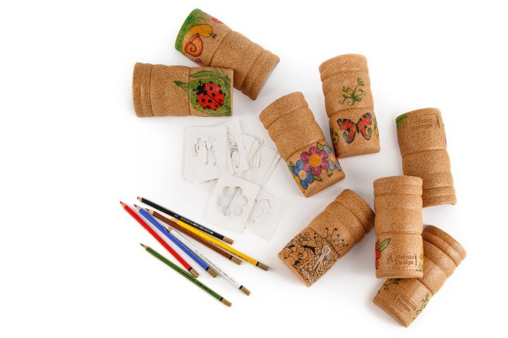 Mehrere braune Vitalisierungsflaschen aus Kork mit bunten, kindgerechten Zeichnungen wie Schmetterlingen, Blumen und Marienkäfern. Daneben liegen Bleistifte, Pinsel, Filzstifte und verschiedene Schablonen mit Motiven.