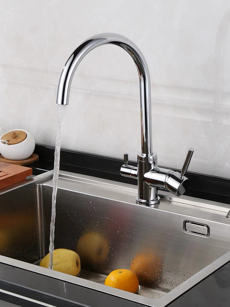 Ein moderner, glänzender Wasserhahn mit hohem Bogen über einem Edelstahl-Spülbecken. Wasser fließt aus dem Hahn. Im Spülbecken liegen drei Früchte, vermutlich Äpfel oder Orangen. Im Hintergrund ist eine helle Wandfliese zu sehen. Links neben der Spüle steht ein rundes Gefäß mit Holzdeckel auf einem Holzbrett. Am rechten Rand des Bildes ist ein Teil eines Bedienhebels des Wasserhahns sichtbar. Die Umgebung wirkt sauber und modern. Die Arbeitsplatte unter dem Spülbecken ist dunkel.