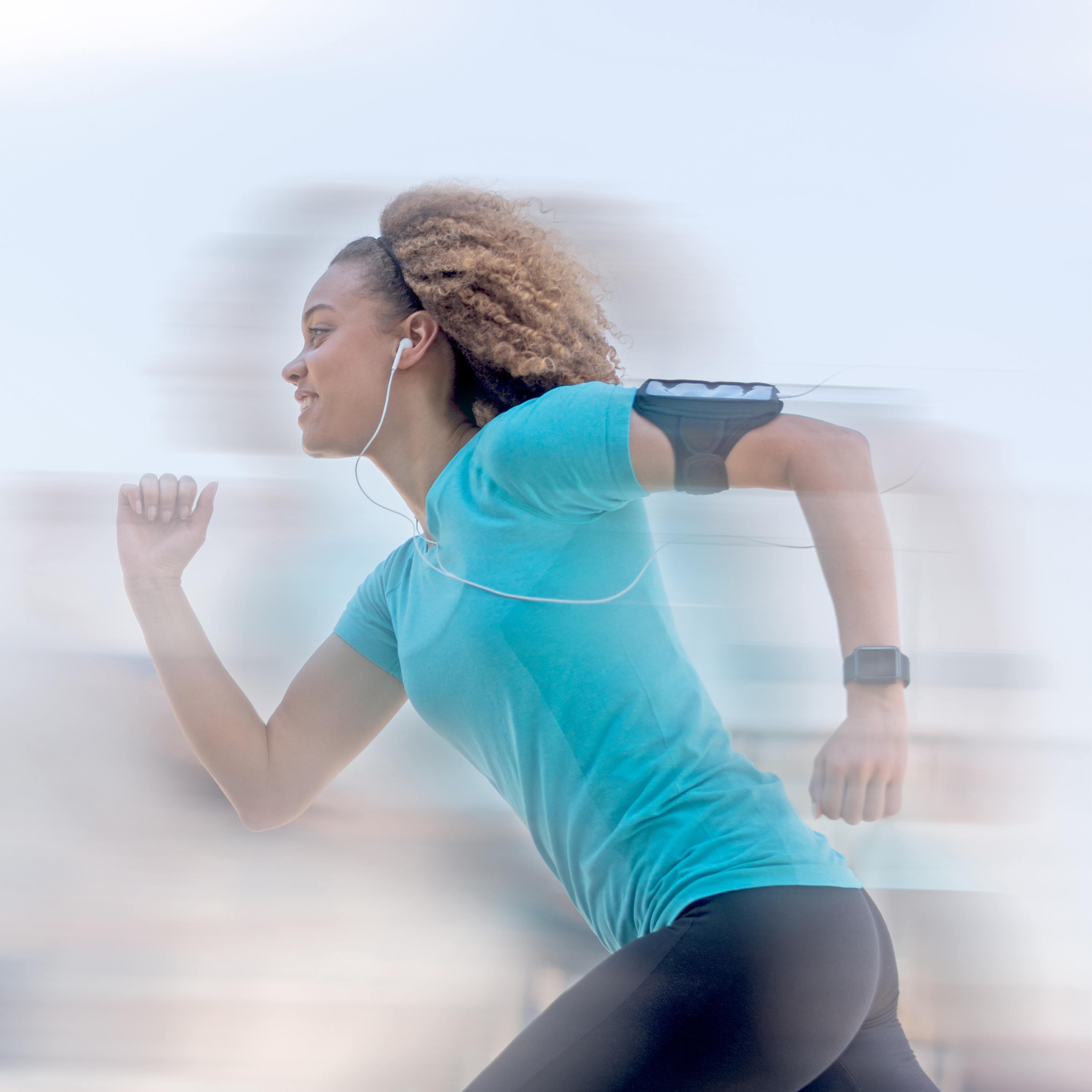 Eine Frau joggt draußen, trägt ein türkisfarbenes Sportshirt, schwarze Leggings, Fitnessarmband, Kopfhörer und eine Armtasche.