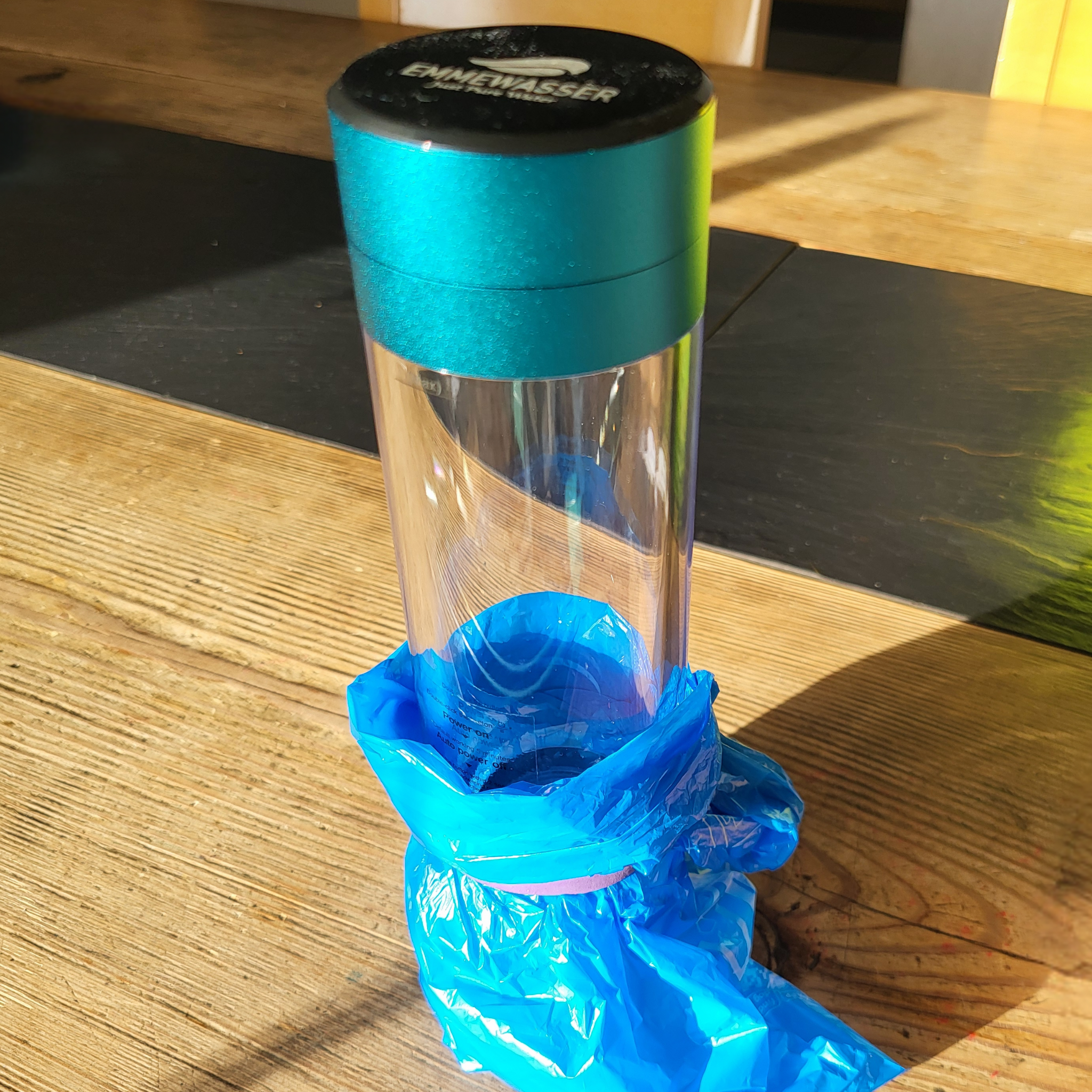 Auf einem Holztisch steht eine transparente Trinkflasche mit türkisfarbenem Schraubdeckel, unten ist eine blaue Plastiktüte befestigt.