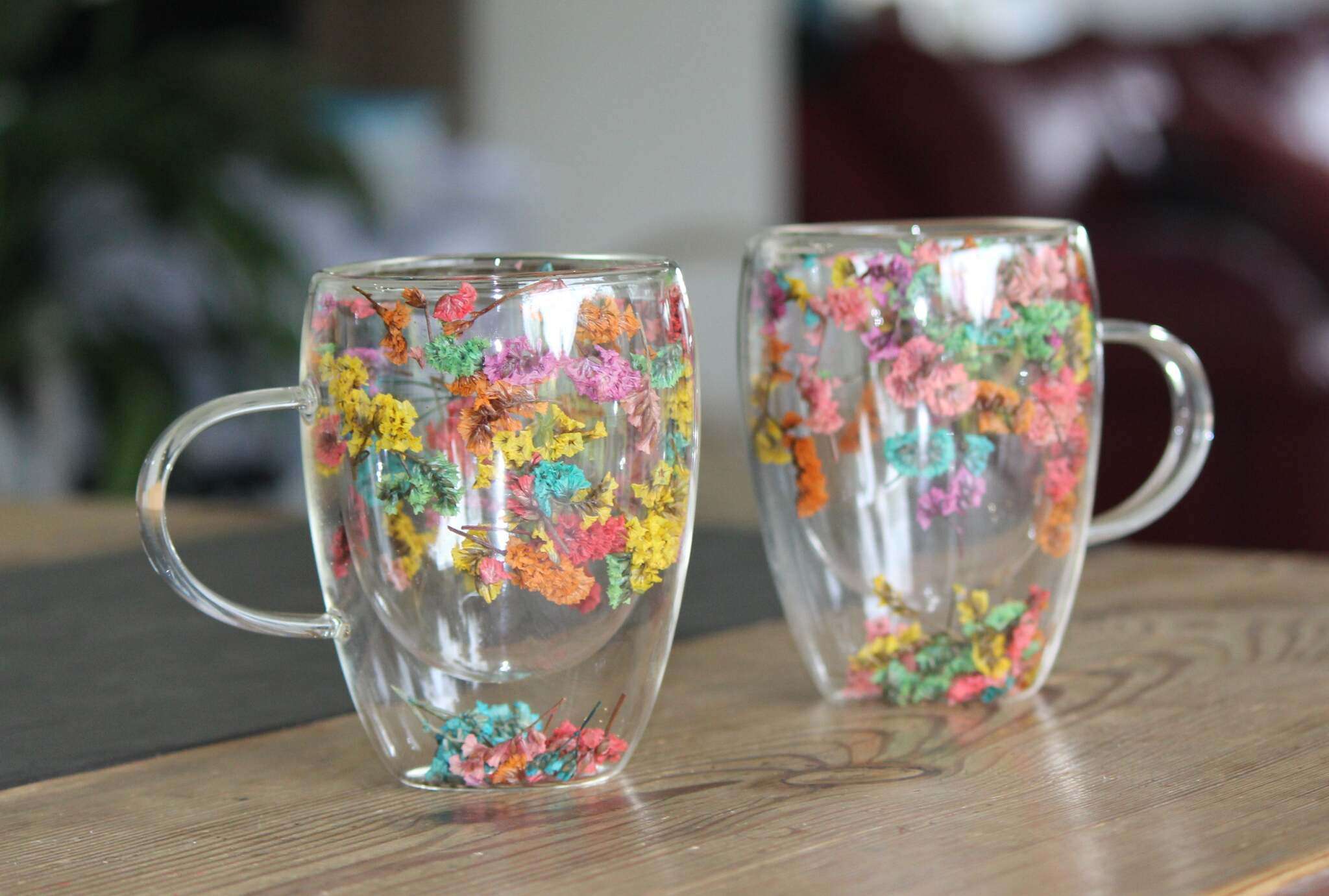 Zwei doppelwandige Glastassen mit Henkeln, verziert mit echten getrockneten Blumen in bunten Farben, auf Holztisch stehend.
