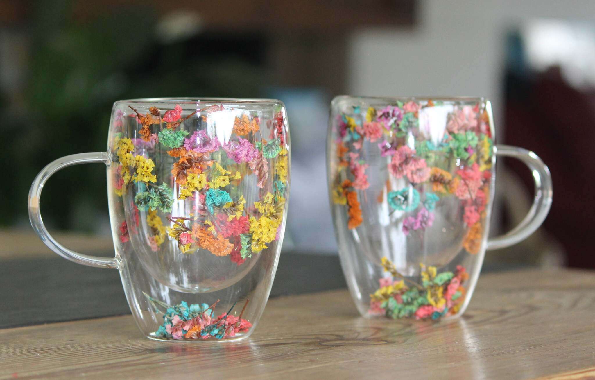 Zwei doppelwandige Glastassen mit Henkeln stehen auf einem Holztisch. Beide Becher sind mit echten, bunten, getrockneten Blumen dekoriert. Die Blumen befinden sich zwischen den Glaswänden und zeigen Farben wie Gelb, Orange, Rot, Grün, Türkis und Lila. Die Tassen sind durchsichtig und reflektieren das Licht. Im Hintergrund sind unscharfe, neutrale Raumdetails zu erkennen.