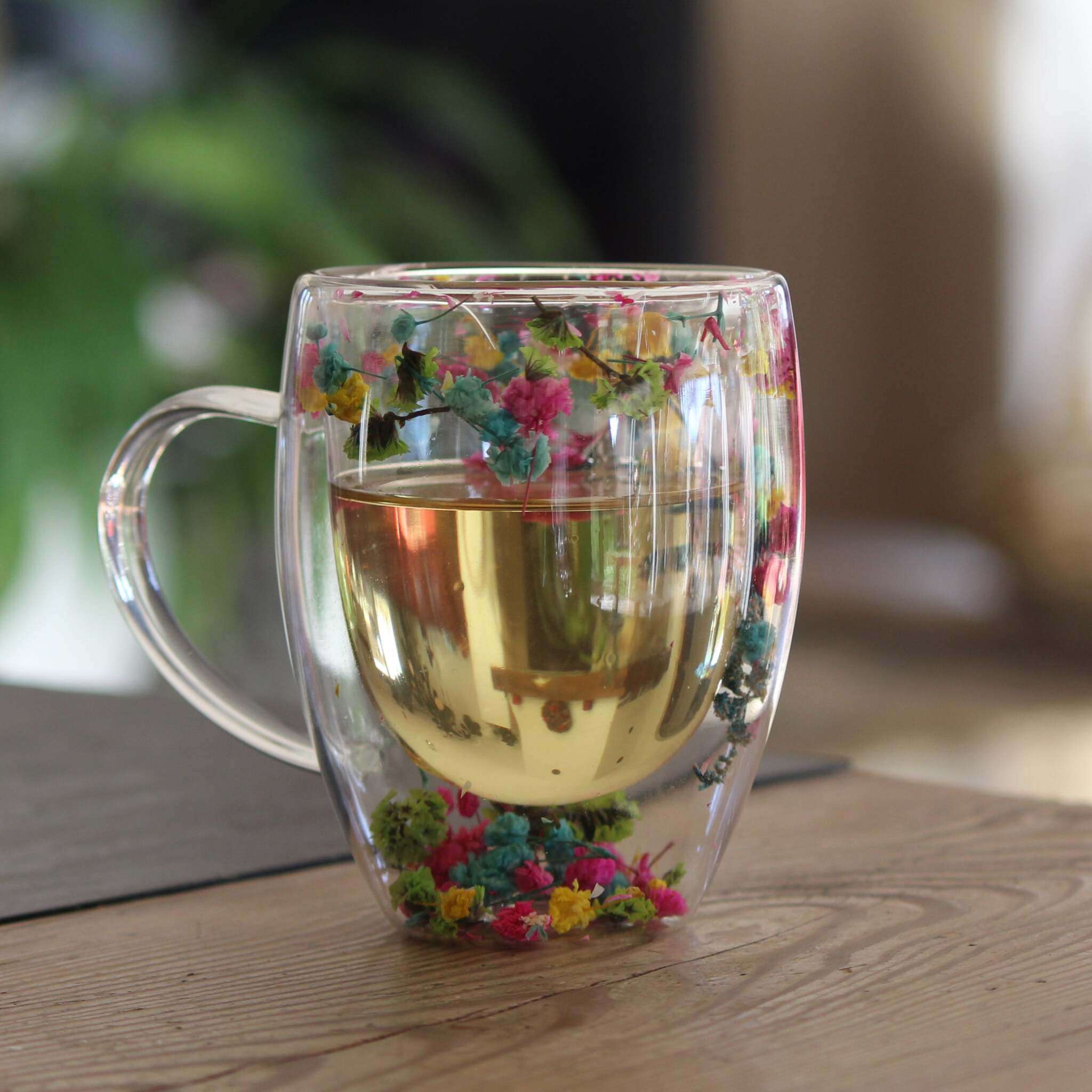 Ein doppelwandiger Glasbecher mit Henkel, gefüllt mit hellem Getränk, dekoriert mit echten getrockneten bunten Blumen.