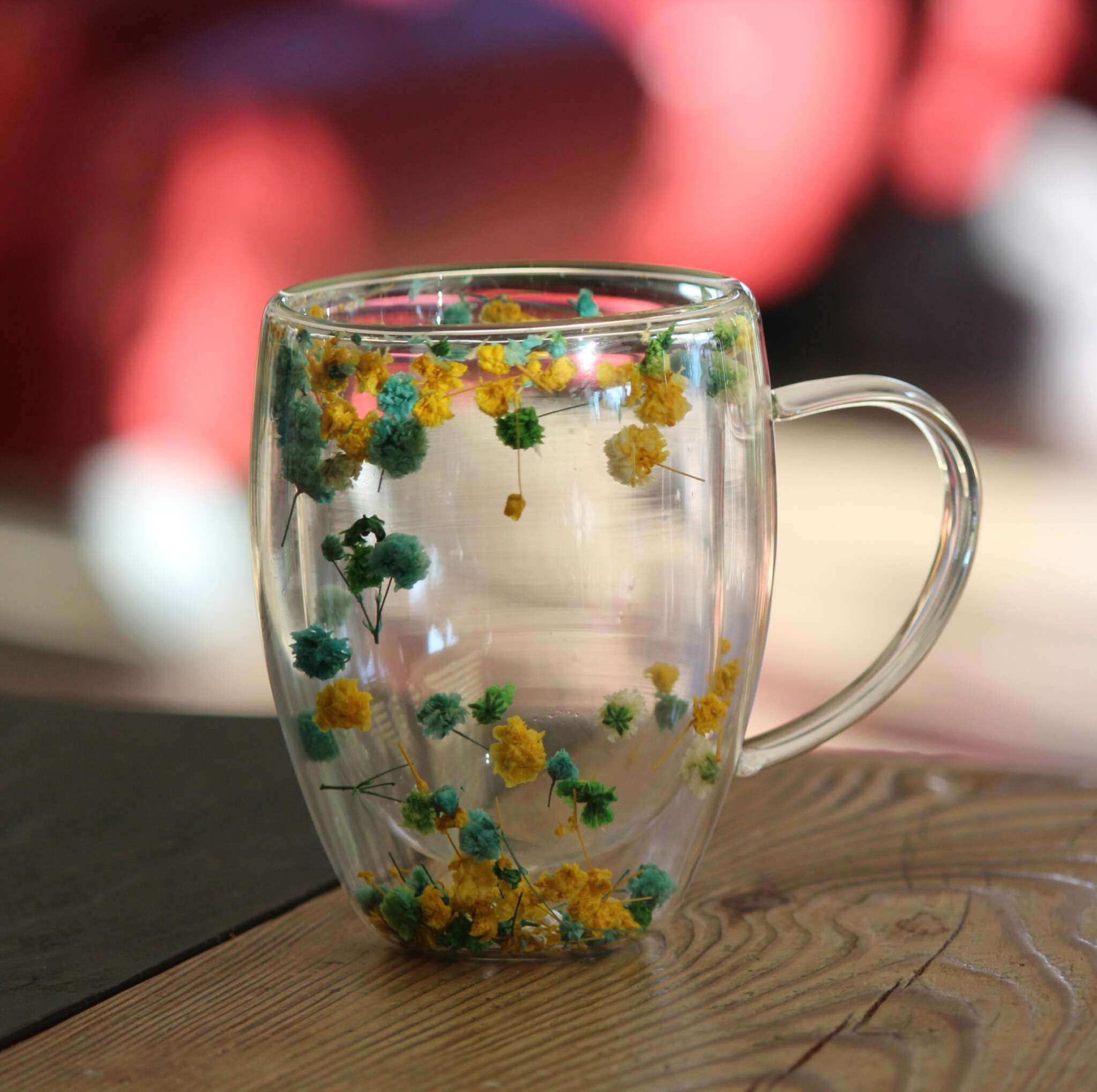 Ein doppelwandiger Glasbecher mit Henkel, verziert mit echten getrockneten gelben und grünen Blumen, auf Holztisch stehend.