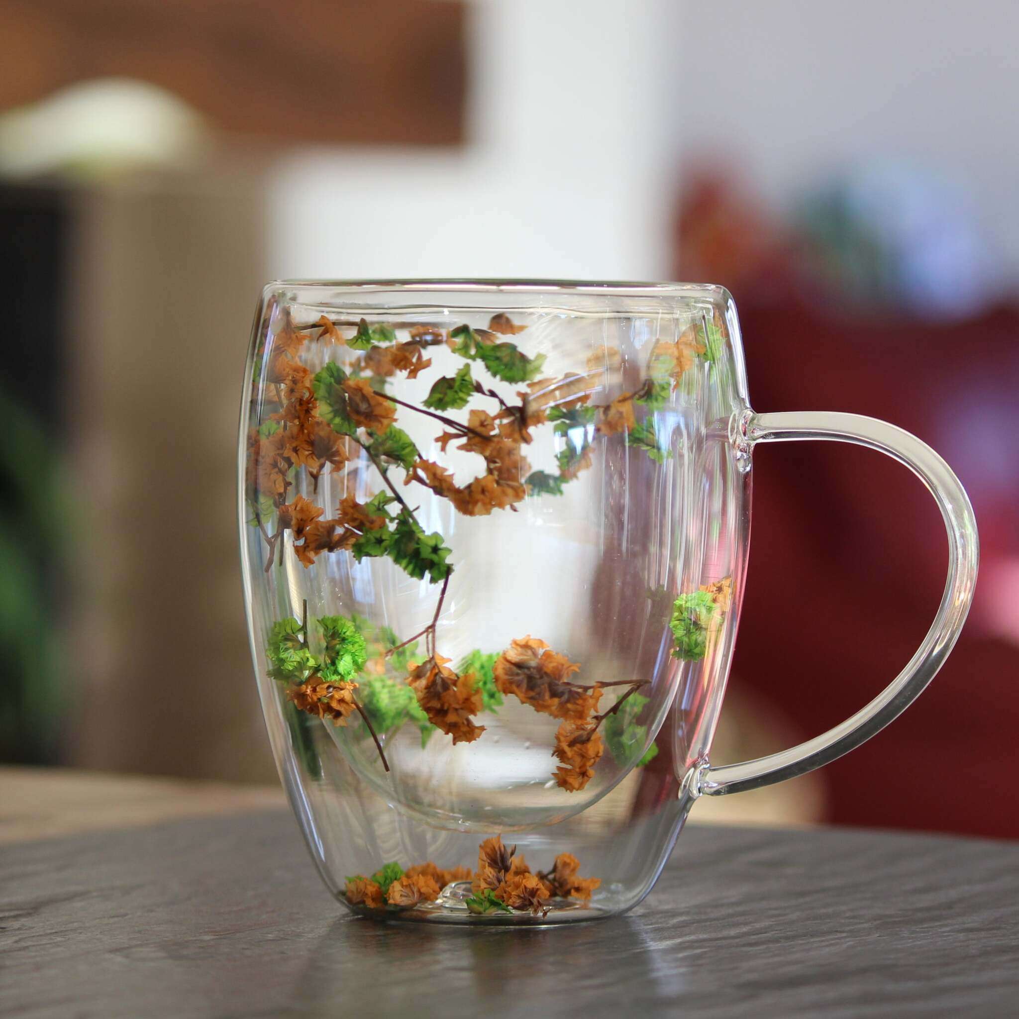 Doppelwandiger Glasbecher mit Henkel, verziert mit echten getrockneten Blumen in Orange und Grün, auf Holztisch stehend.