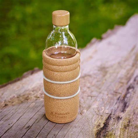 Eine Glas-Trinkflasche mit rundem Holzdeckel und abnehmbarer Korkhülle steht auf einem verwitterten Baumstamm im Freien.
