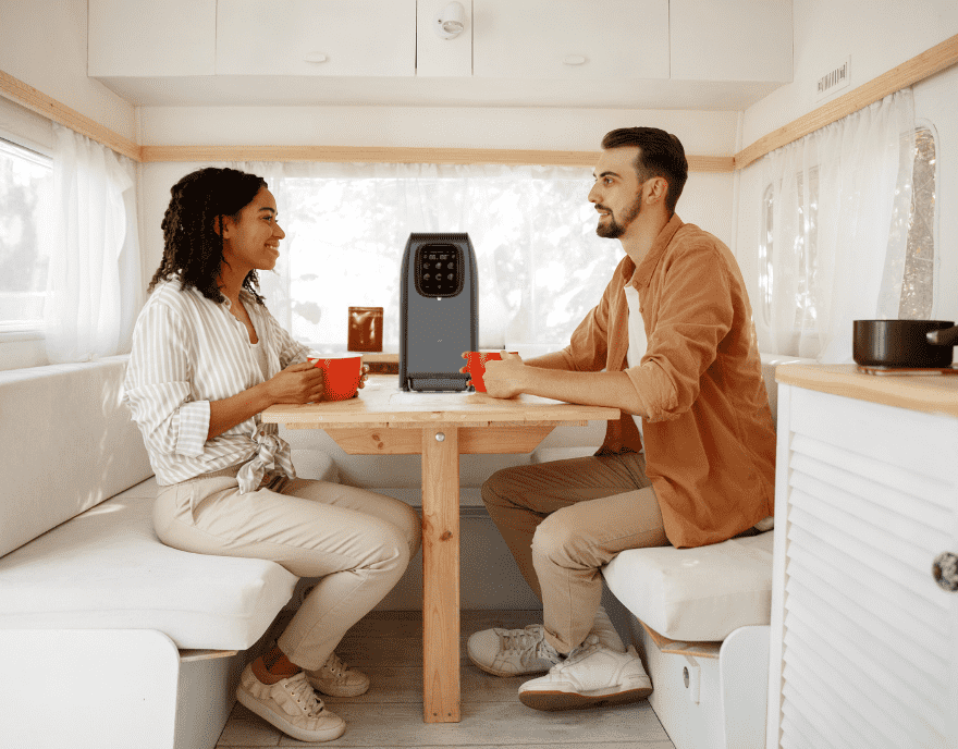 Zwei Personen sitzen in einem hellen Wohnmobil an einem Holztisch, trinken aus roten Tassen, tragbare Powerstation auf dem Tisch.