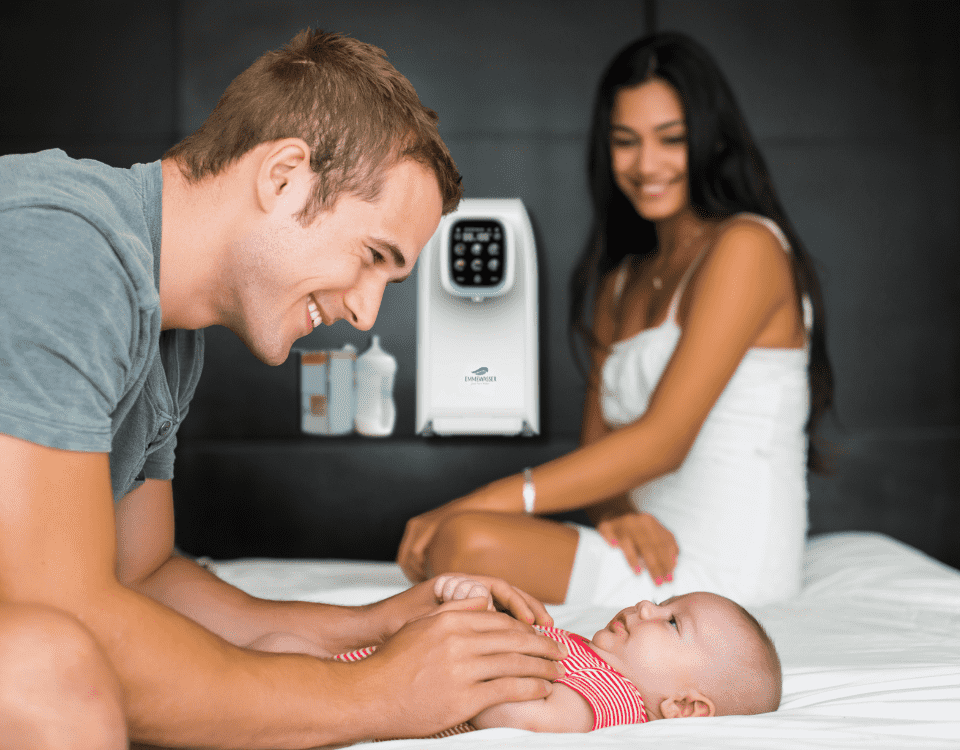Ein lächelnder Mann spielt mit einem Baby auf einem Bett, eine Frau sitzt im Hintergrund, auf einem Regal steht ein Babyflaschenzubereiter.