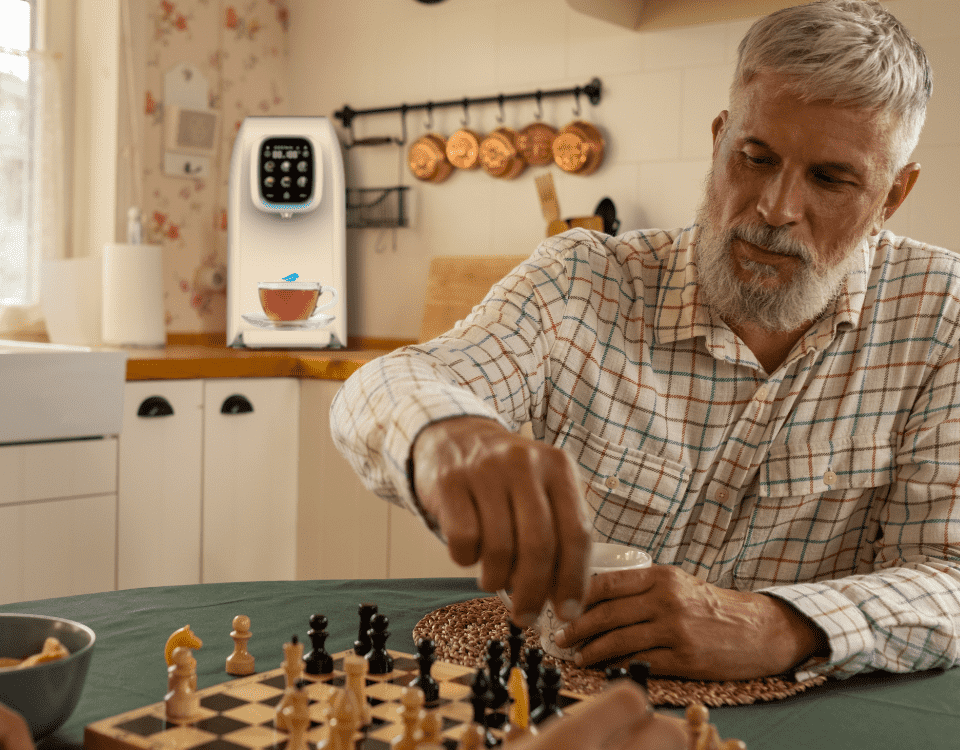 Ein älterer Mann mit grauem Bart spielt Schach an einem Holztisch in einer wohnlichen Küche, hält eine Tasse, im Hintergrund eine moderne Kaffeemaschine sowie Kupfertöpfe an der Wand.