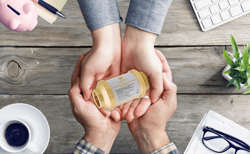 Zwei paar Hände, ein größeres hält ein kleineres, gemeinsam einen goldfarbenen UMH Wasserwirbler auf Holztisch haltend. Daneben eine Kaffeetasse, grüne Pflanze, Tastatur, Brille, rosa Sparschwein und Stift sichtbar.