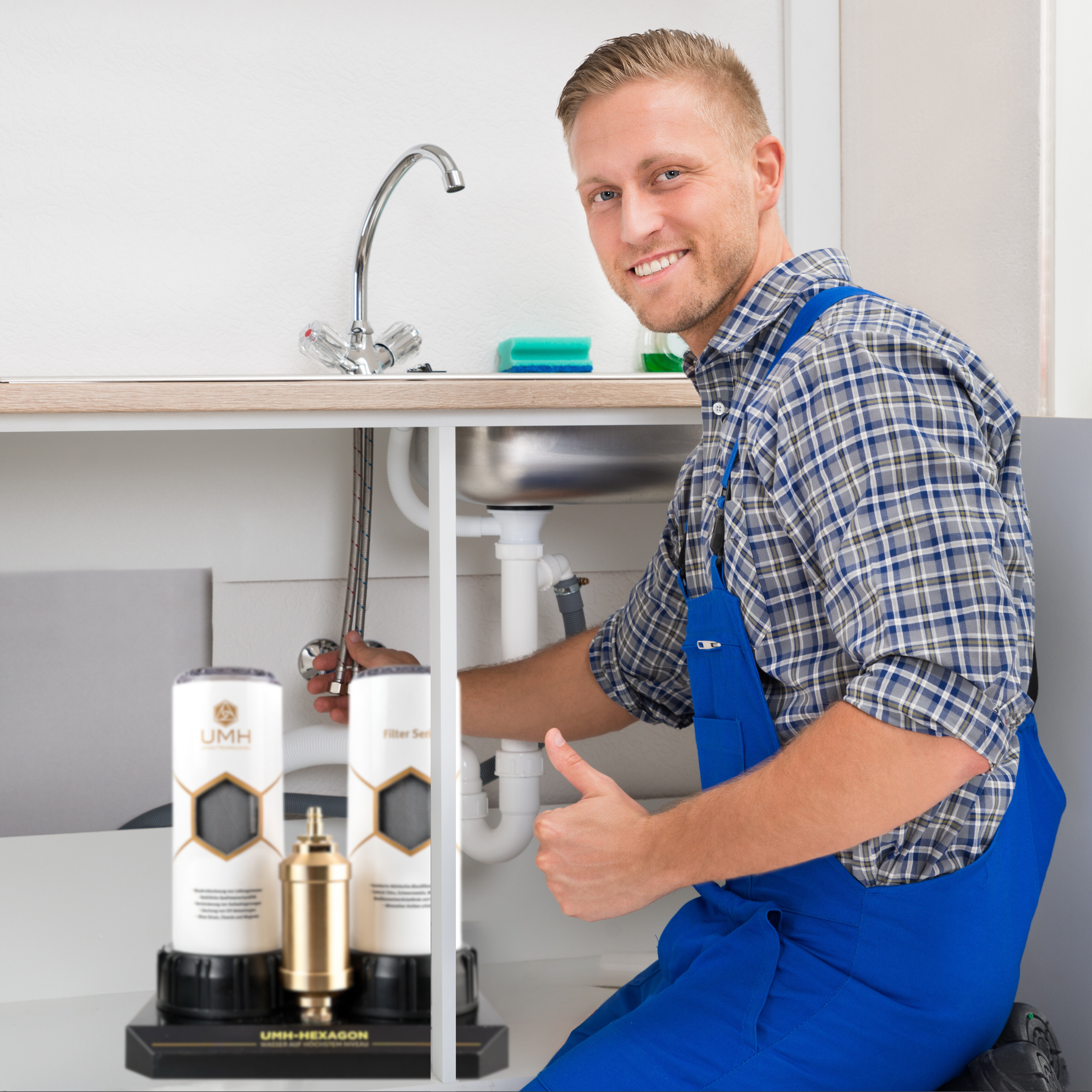 Ein Handwerker in blauer Latzhose arbeitet unter einem Waschbecken an Rohren, daneben steht ein Wasserfilter-Set von UMH.
