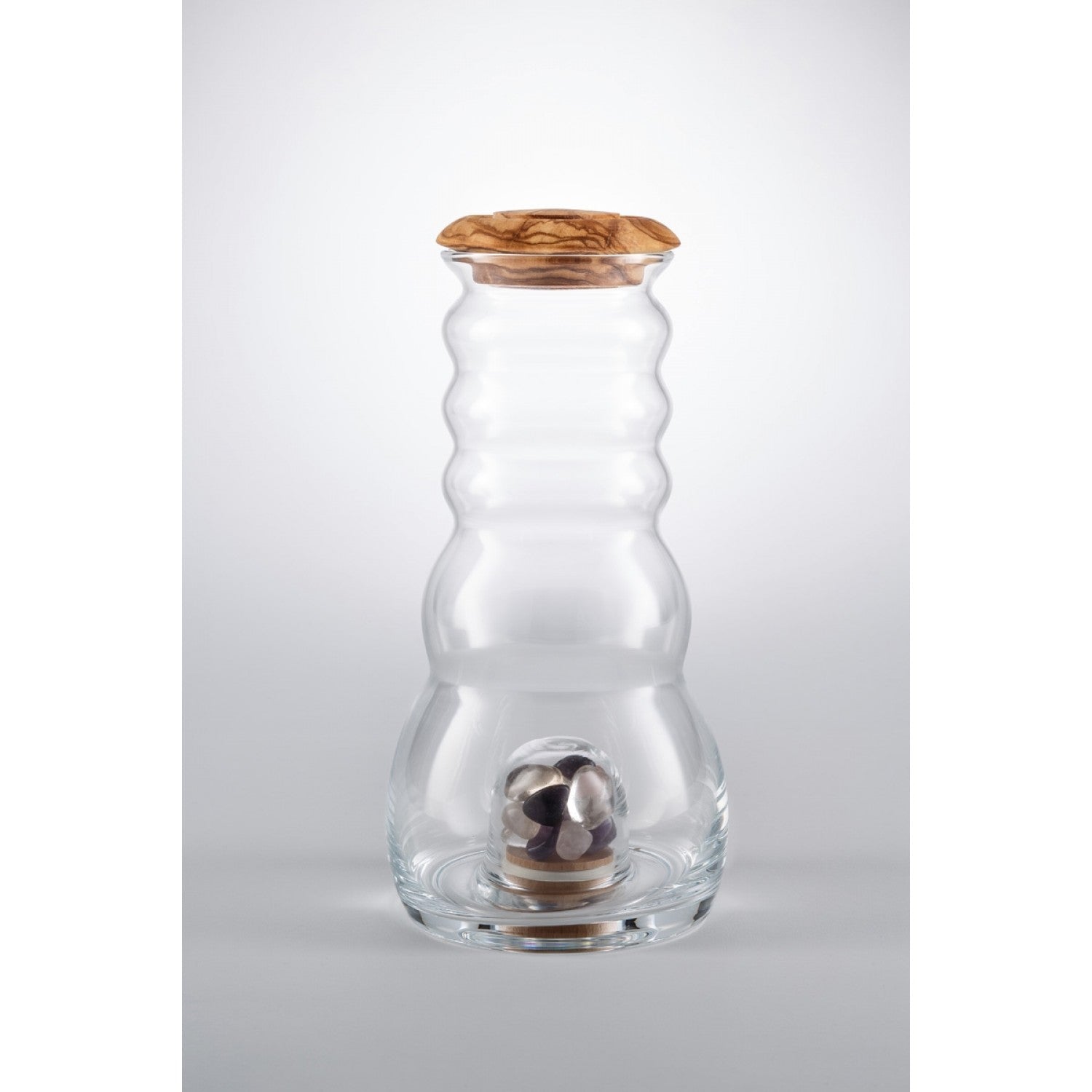 Eine klare Cadus Karaffe 1 Liter mit gewelltem Glasdesign, Holzdeckel und Glasfach im Boden, gefüllt mit Edelsteinen.