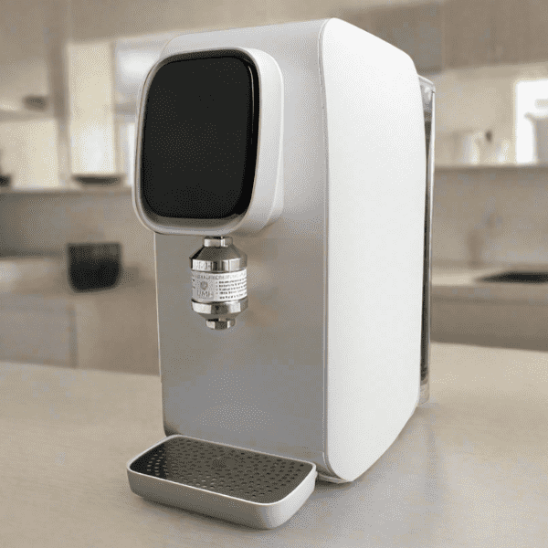 Auf einer Küchenarbeitsplatte steht ein moderner, weiß-silberner Wasserenergetisierer mit rechteckigem Gehäuse. An der Vorderseite ist ein schwarzes Bedienfeld und ein Metalleinsatz, darunter befindet sich ein kleiner silberner Wasserhahnaufsatz mit Beschriftung sowie eine abnehmbare Abtropfschale aus Metall. Im Hintergrund sind unscharf Küchenschränke, ein Fenster und weitere Küchengeräte zu sehen.