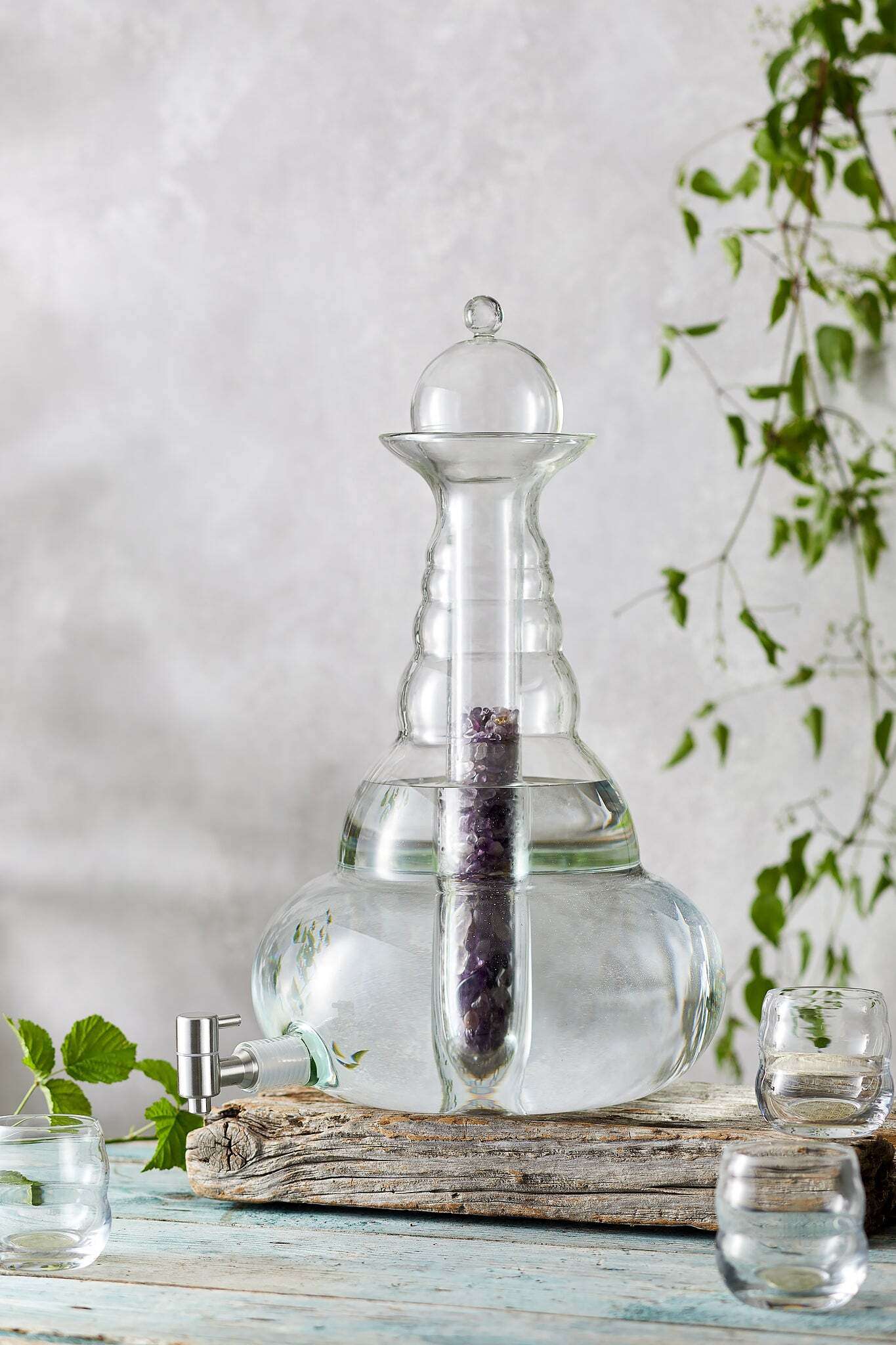 Eine große, bauchige Glaskaraffe mit Deckel und Edelstahlhahn, gefüllt mit Wasser, auf Holzbrettern, umgeben von Gläsern.