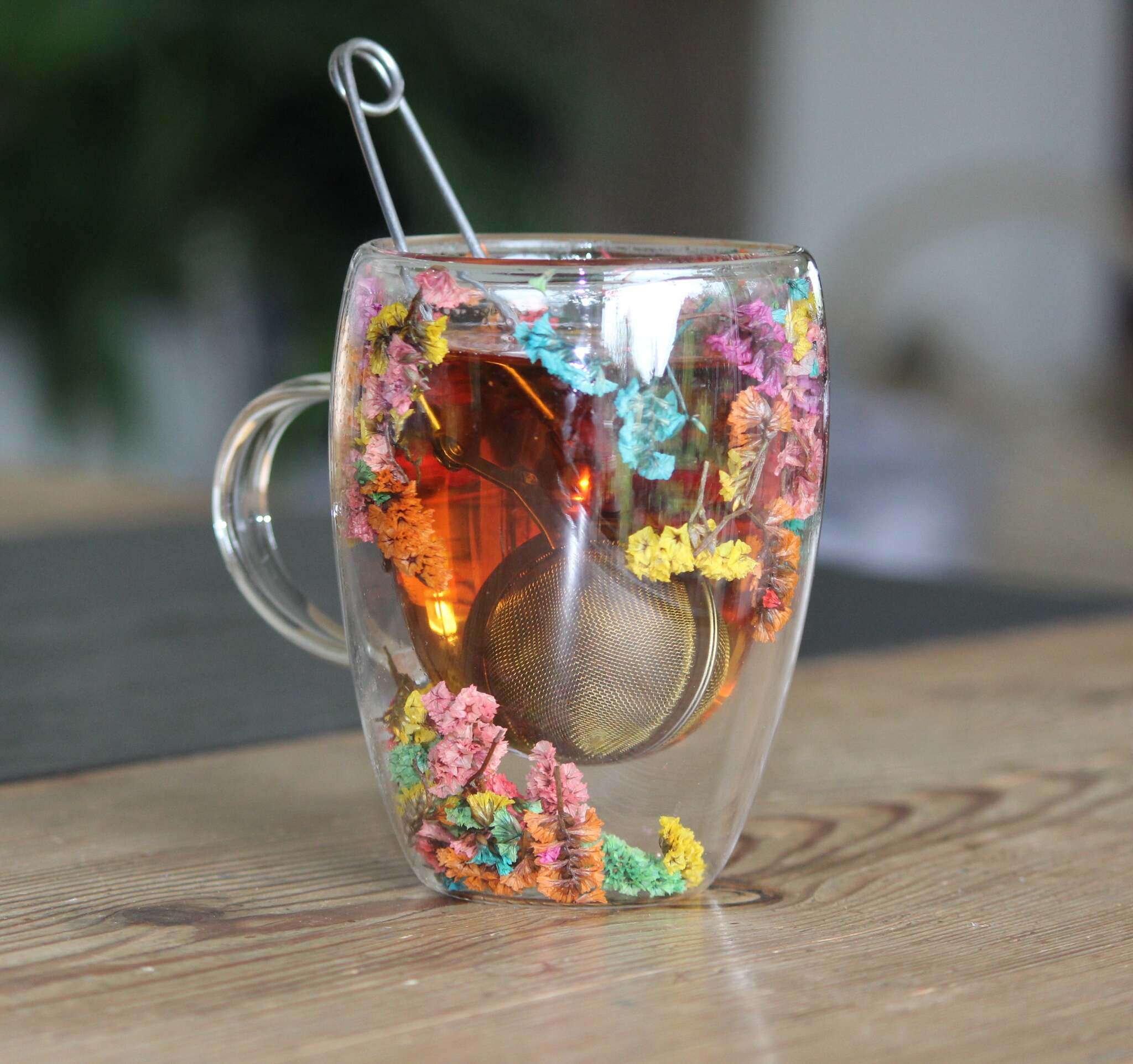 Ein doppelwandiger Glasbecher mit Henkel, dekoriert mit echten getrockneten bunten Blumen, enthält Tee und ein Teesieb.